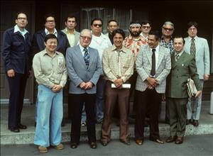 Fourteen men, some Native American, some white, and one Asian, pose for the camera in two rows. Some are wearing bell bottoms. 